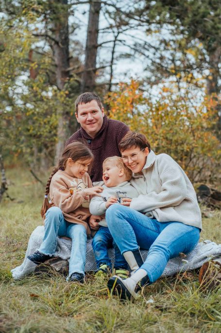Eine Familie mit zwei Erwachsenen und zwei Kindern sitzt fröhlich im Freien unter Bäumen.