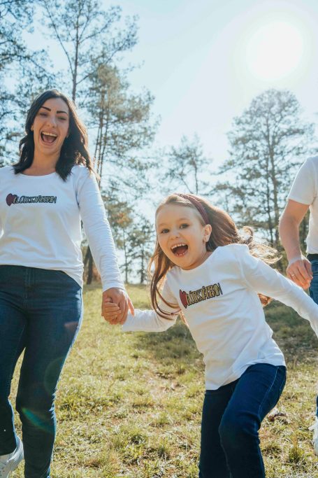 Frau und Mädchen halten sich an den Händen und lachen im Freien unter Sonnenlicht.