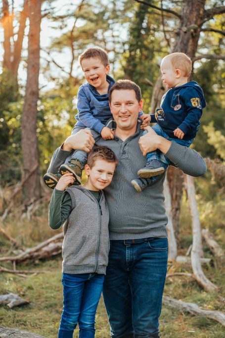Ein Mann mit drei fröhlichen Kindern im Wald, zwei Kinder auf seinen Armen.