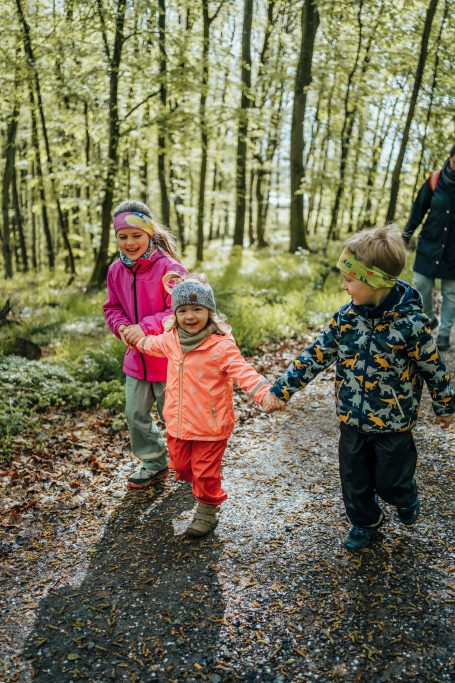 Drei Kinder in bunten Jacken gehen vergnügt in einem Wald.
