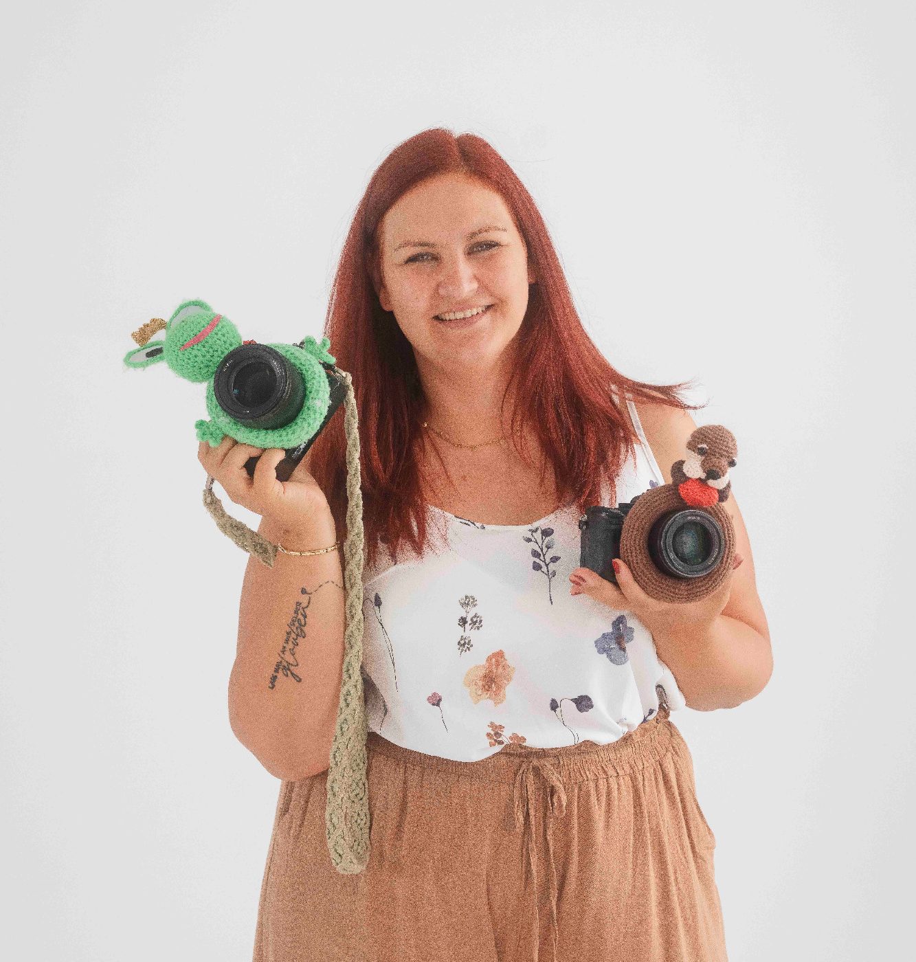 Frau mit rotem Haar hält zwei Kameras in den Händen, lächelt freundlich. Auf den Kameras sitzt ein gehäkelter Frosch und ein Otter.