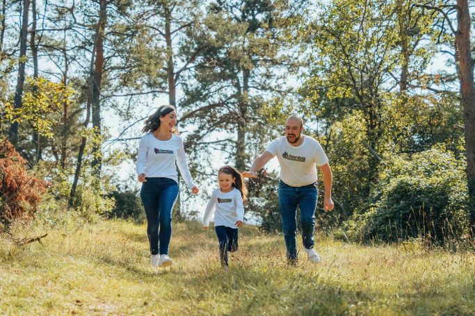 Eine Familie mit einem Kind läuft fröhlich durch einen bewaldeten Weg.