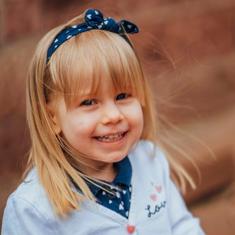 Lächelndes blondes Mädchen mit Haarband, trägt einen hellblauen Pullover.