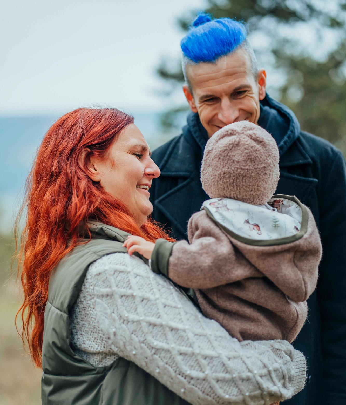 Familie im Freien: Eine Frau mit roten Haaren, ein Mann mit blauen Haaren und ein Baby.