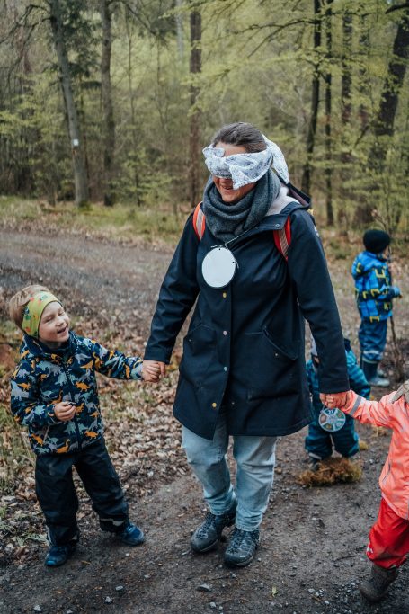 Eine Frau mit Augenbinde hält die Hände von zwei Kindern beim Spazieren im Wald.
