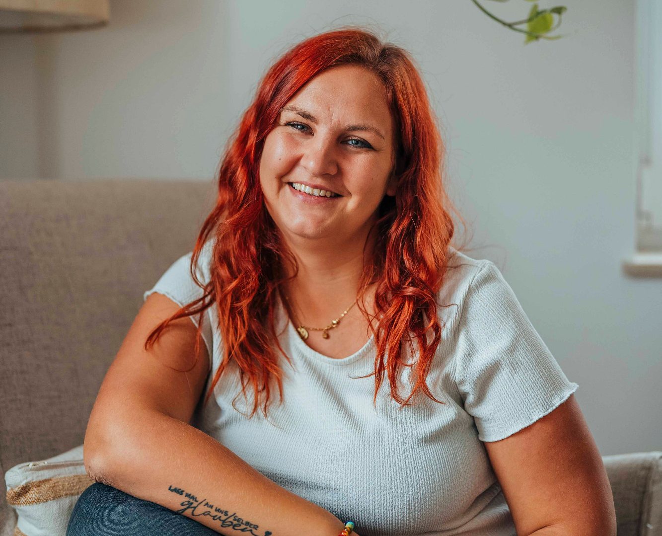 Lächelnde Frau mit langen roten Haaren sitzt auf einem Sessel und trägt ein helles T-Shirt.