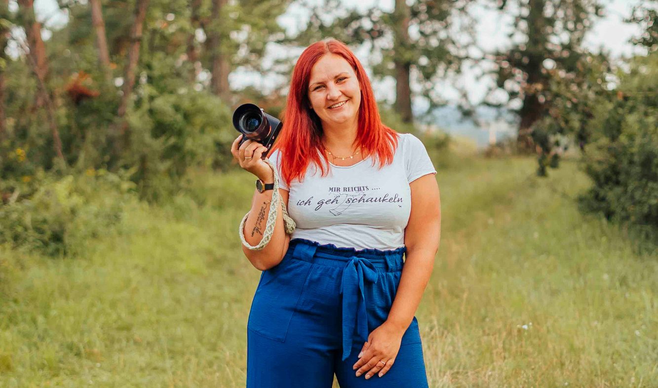Lächelnde Frau mit rotem Haar hält eine Kamera in der Natur.
