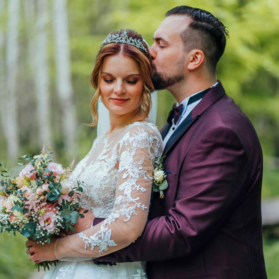 Braut und Bräutigam umarmen sich im Freien, umgeben von Bäumen und Blumen.
