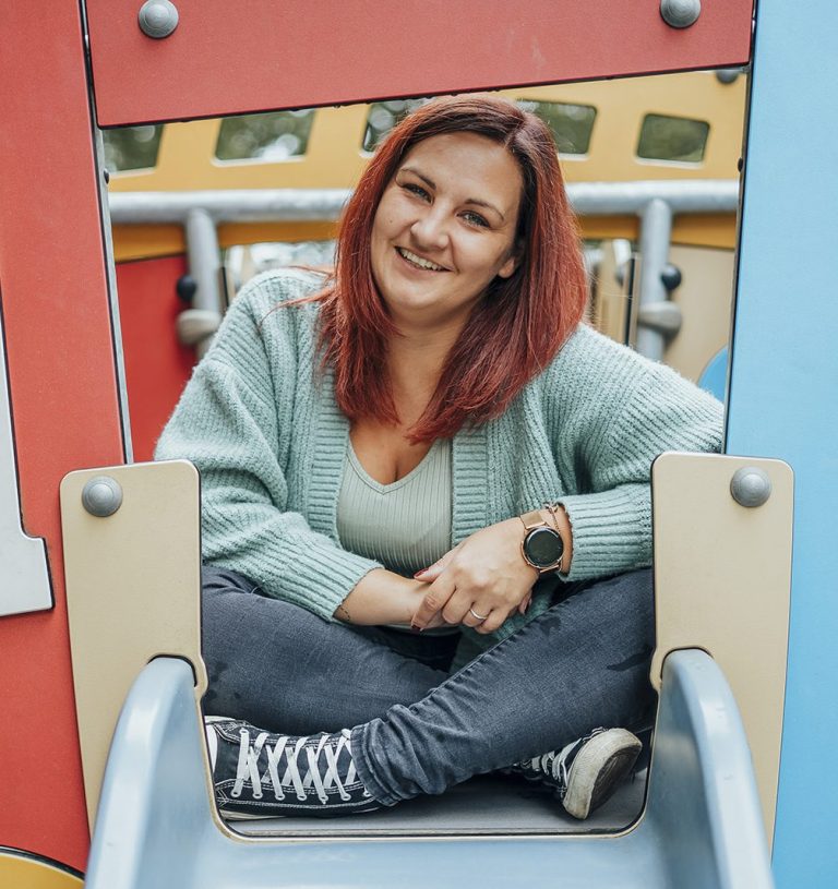 Lachende Frau mit rotem Haar sitzt auf einer Rutsche im Spielplatzbereich.