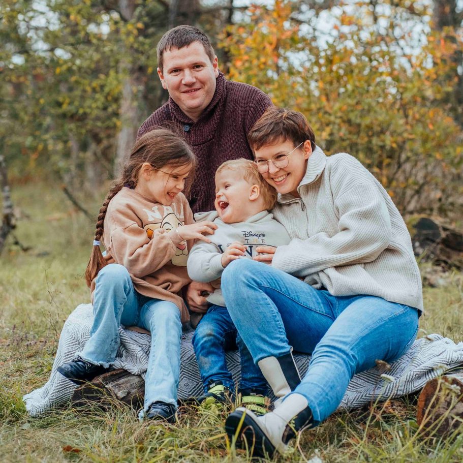Familie mit zwei Kindern sitzt fröhlich im Freien auf einem Baumstamm, umgeben von Natur.