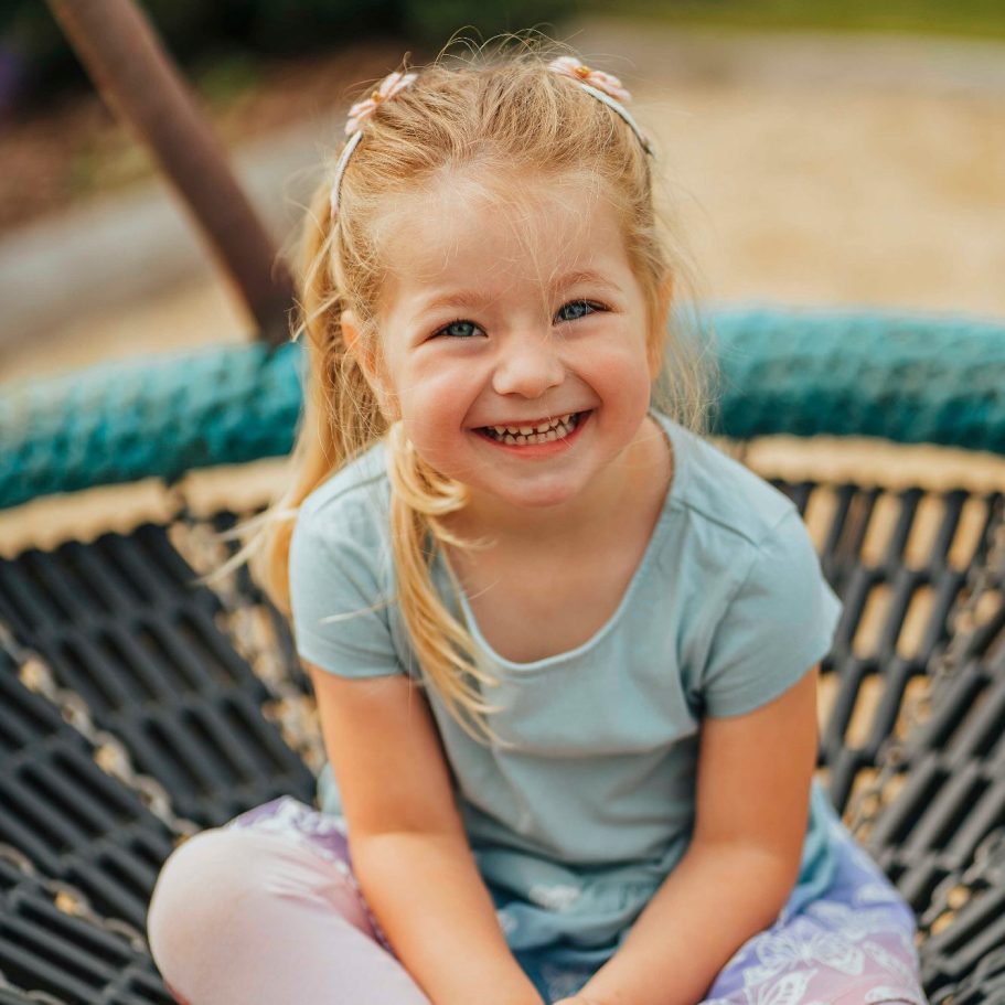 Ein lächelndes Mädchen sitzt auf einer Schaukel und trägt ein hellblaues T-Shirt.