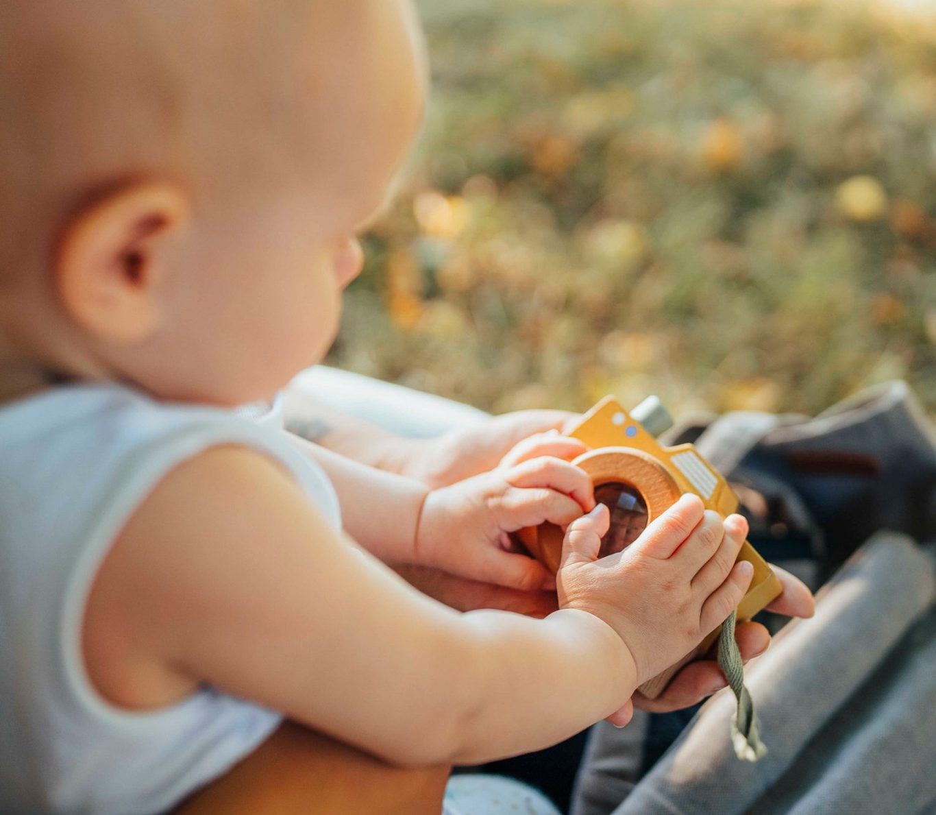 Ein Baby hält ein orangefarbenes Spielzeugkamera aus Holz im Freien.
