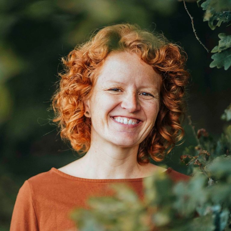 Lächelnde Frau mit roten lockigen Haaren steht vor grünem Hintergrund.