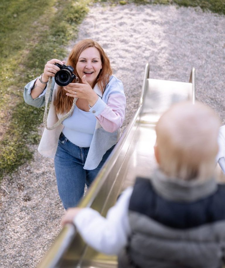 Frau mit Kamera lächelt, während ein Kleinkind auf einer Rutsche sitzt.