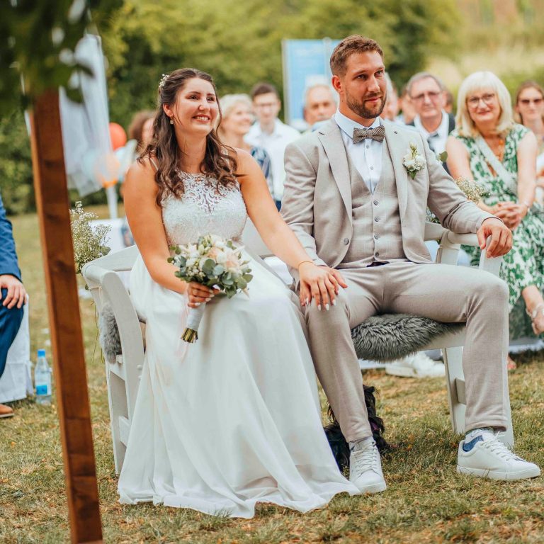 Braut und Bräutigam sitzen gemeinsam, umgeben von Gästen in festlicher Atmosphäre.