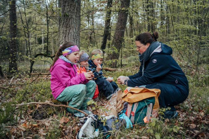 Eine Frau sitzt mit drei Kindern im Wald und genießt einen Snack in der Natur.