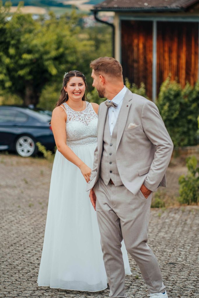 Braut und Bräutigam stehen lächelnd im Freien, umgeben von grüner Vegetation.