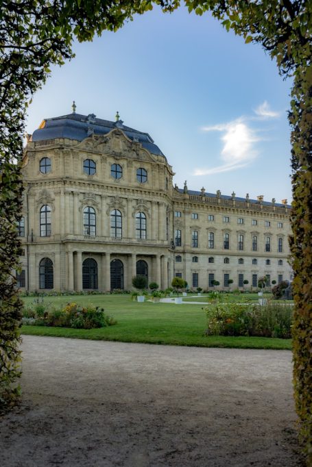 Barockes Schloss mit gepflegtem Garten, umgeben von Bäumen und blauen Himmel.