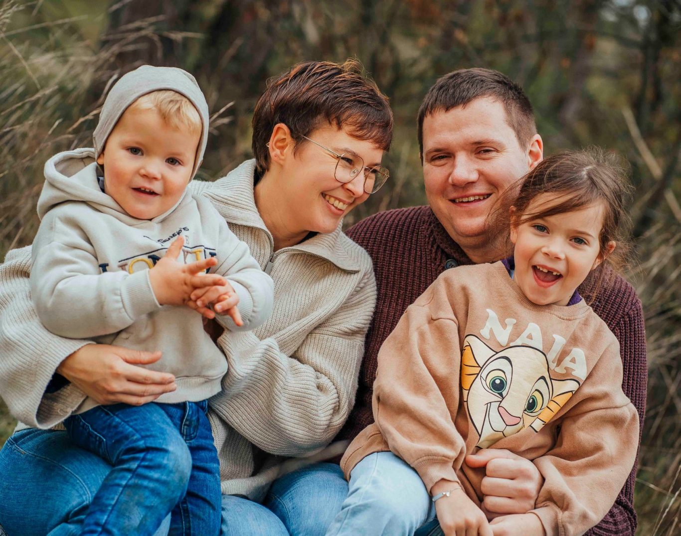 Familie posiert lächelnd im Freien, Kinder in Pullovern, Eltern umarmend.