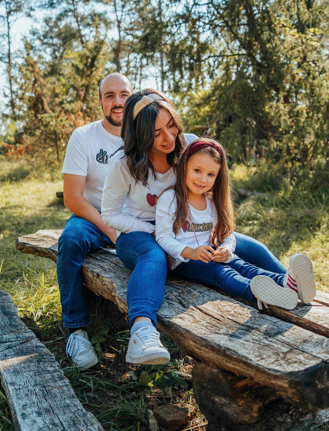 Familie sitzt auf einer Holzbank in der Natur, lächelt in die Kamera.