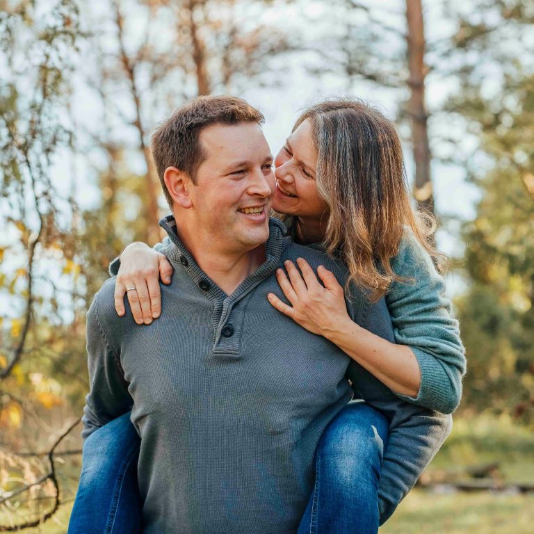 Ein Mann trägt eine Frau auf dem Rücken, beide lachen in einer natürlichen Umgebung.